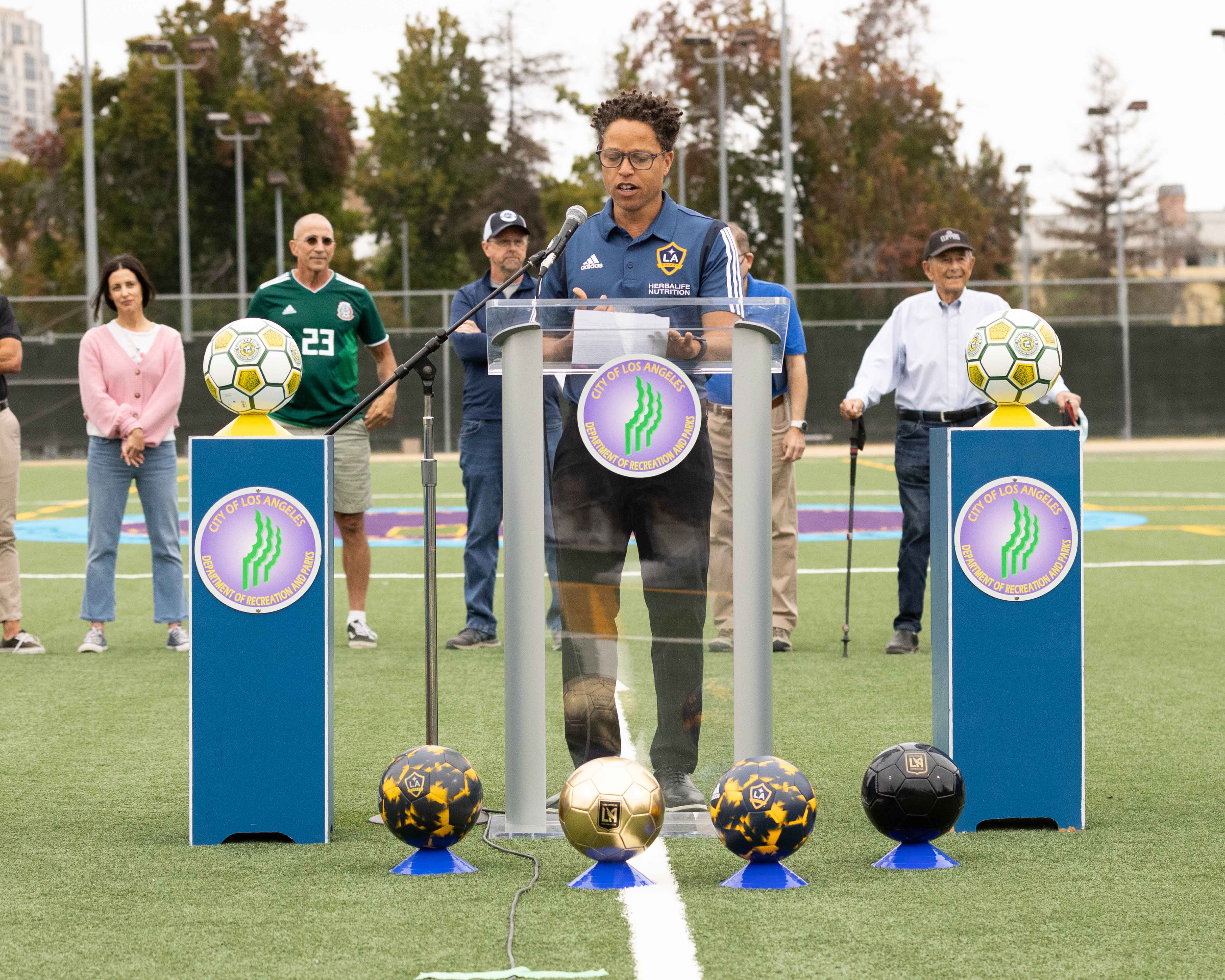 LA Galaxy legend Cobi Jones stands at a podium in the middle of a soccer field to give remarks at the field grand opening. 