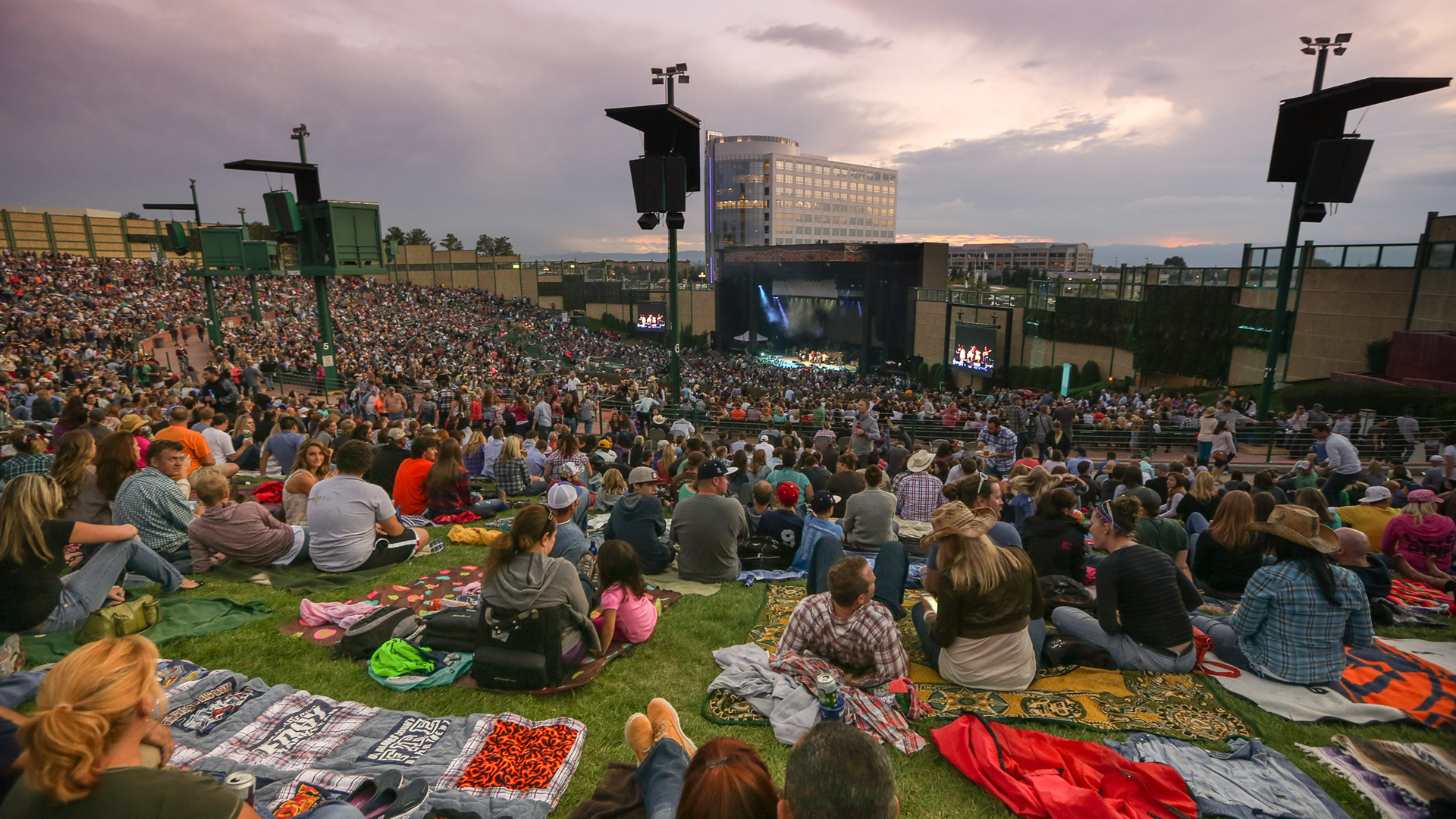 Packed lawn and seats facing the lit stage at dusk