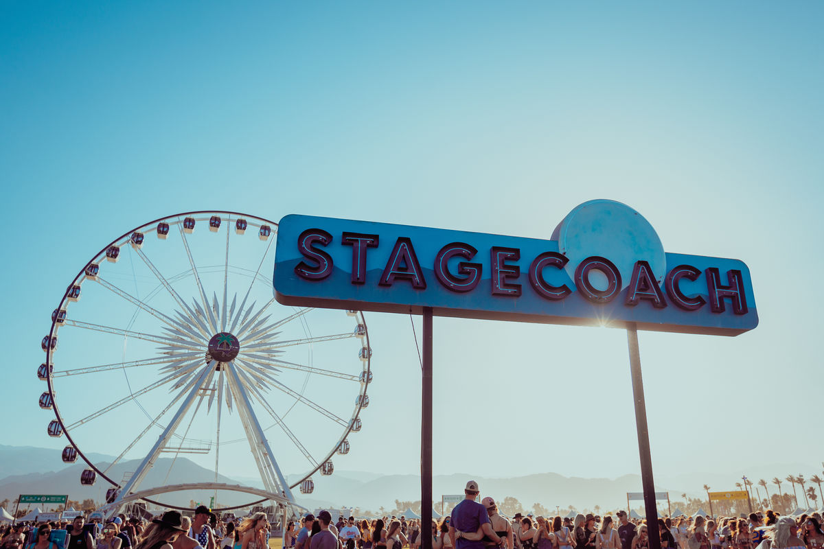 Image from behind of a performer playing the guitar and jumping in the air with the crowd and ferris wheel in front of him at Stagecoach