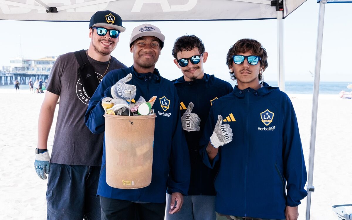 Four volunteers wear LA Galaxy jackets and gloves while holding a bucket filled with collected trash during a beach cleanup.