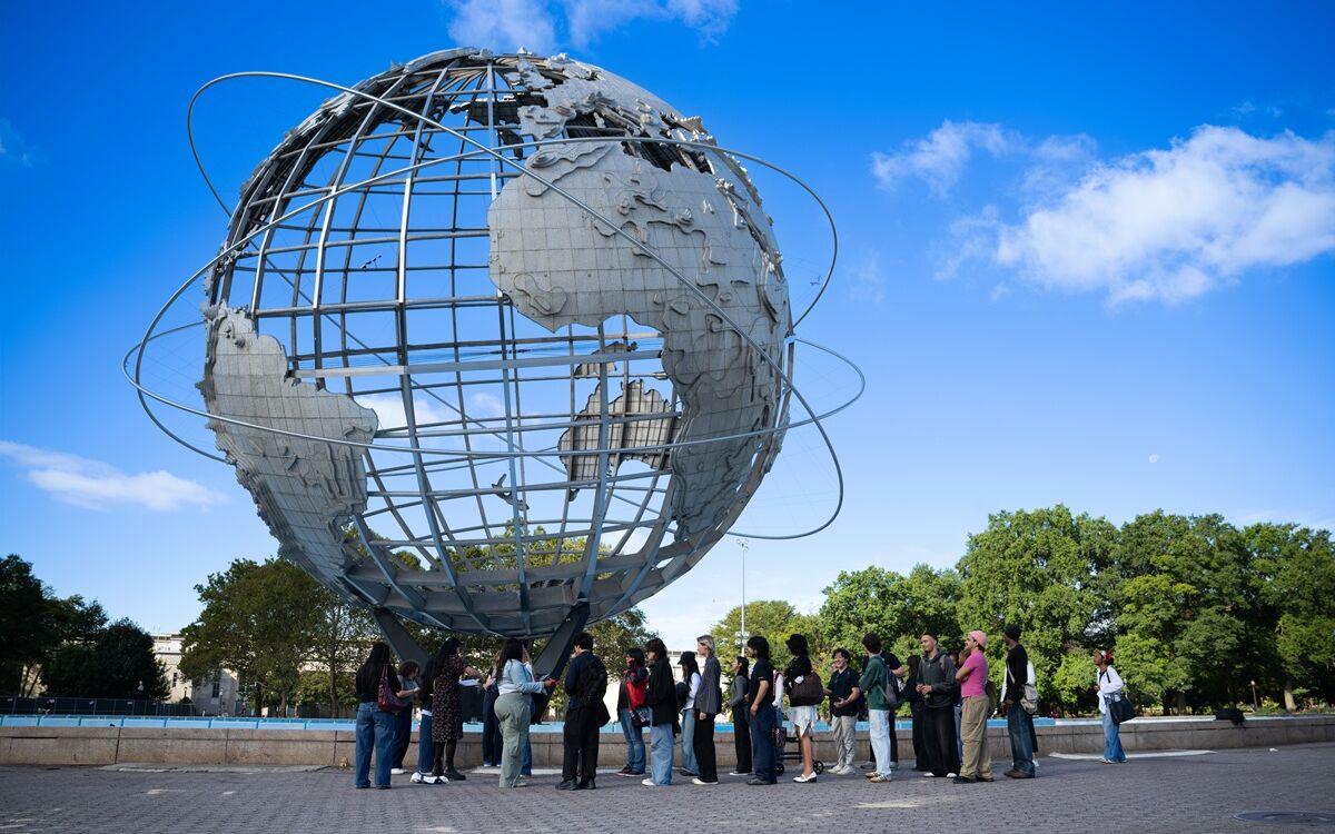 Group gathers outdoors beneath the Unisphere at Flushing Meadows Corona Park under a bright blue sky.