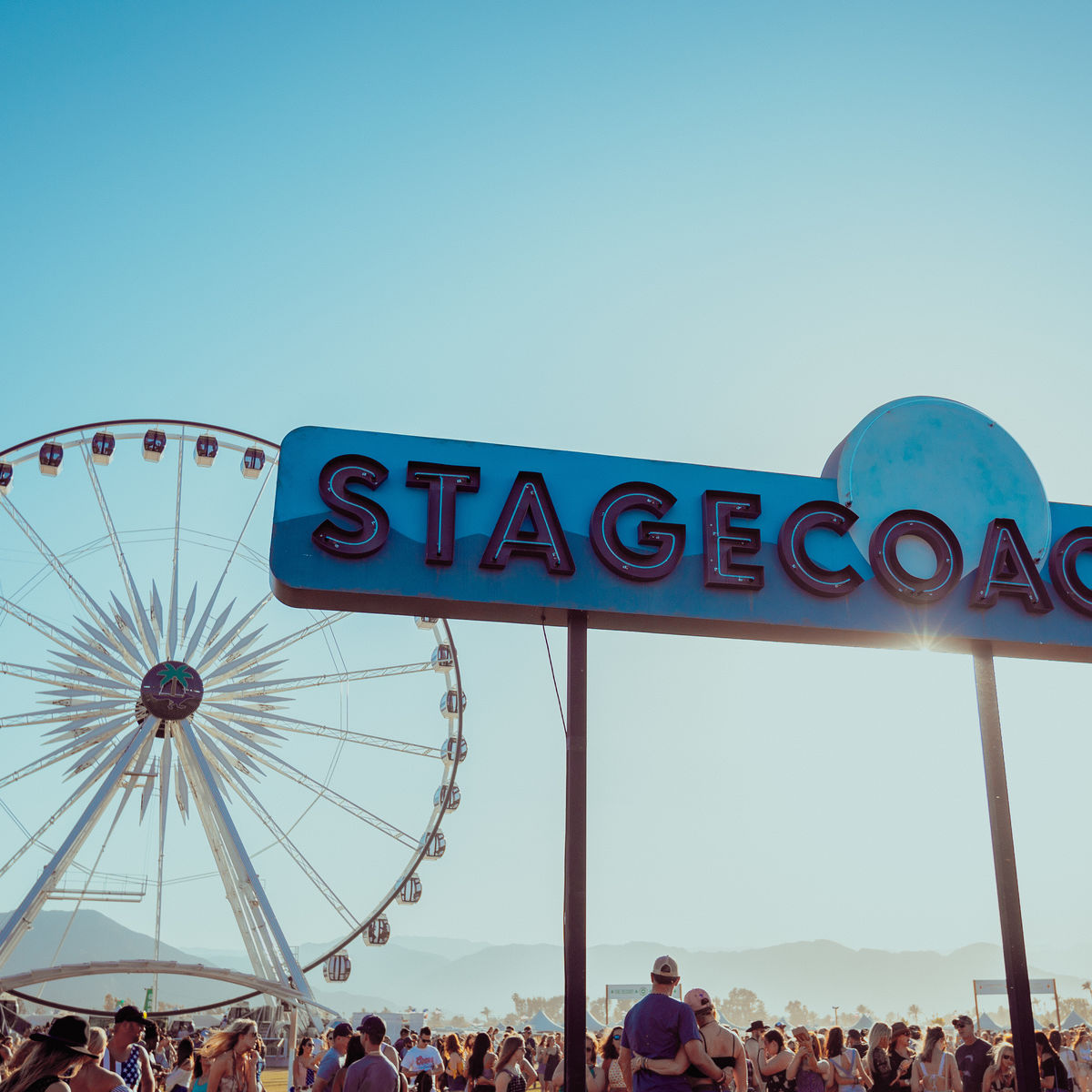 Image from behind of a performer playing the guitar and jumping in the air with the crowd and ferris wheel in front of him at Stagecoach